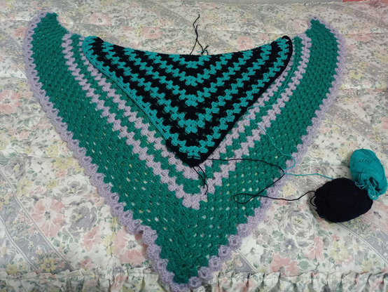 Una colcha floreada, sobre ésta el primer chal que hice y, a su vez, sobre este, el nuevo chal que estoy haciendo, siendo casi la mitad. A la derecha están los dos ovillos que estoy usando, el negro y el verde ópalo.