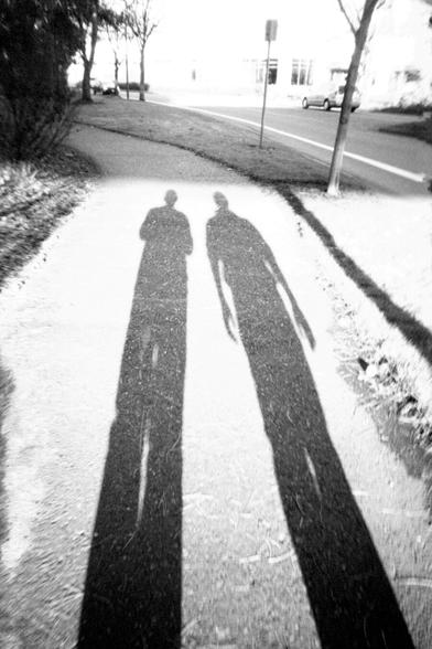 A black and white photo of the elongated shadows of two people being cast on the sidewalk.