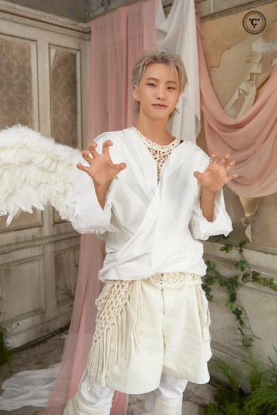 The image features Hoshi wearing a white outfit with angelic wings, posing playfully while making the horanghae gesture. The background includes soft drapes in shades of pink and an ornate, vintage setting.