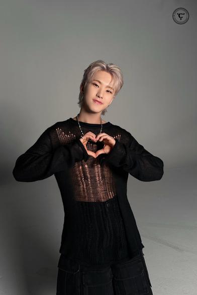Hoshi with light blonde hair is wearing a black knit sweater and black pants, posing by making a heart shape with his hands. The background is a neutral gray.