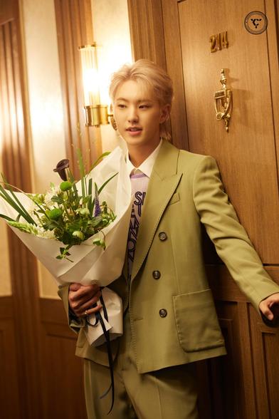 Hoshi in a light green suit holds a bouquet of flowers, standing by a wooden door with the number 214. The setting features soft lighting and a warm wood texture, creating an elegant atmosphere.