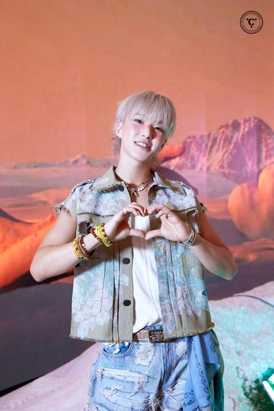 Hoshi with short blond hair is making a heart shape with his hands. He is wearing a floral patterned sleeveless shirt over a white top and distressed denim shorts. The background features a colorful, rocky landscape.