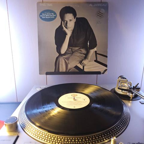 A vinyl record sits on a turntable. Behind the turntable, a vinyl album outer sleeve is displayed. The front cover shows Al Jarreau sitting on the arm of a chair with his chin resting on his palm.