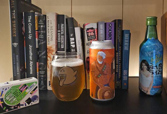 A large stemless wine glass filled with beer and a 16 ounce can sit on a shelf. The glass features art of Boo from Mario Brothers holding a beer. The can art features a shirtless cowboy holding his hat down in front of his pants.

Behind are some books about various music genres (80's rock, hip hop) and artists (2Pac, Aaliyah, Mariah Carey, etc.), as well as a soju bottle featuring Hyori, and a Pocket Rocker.