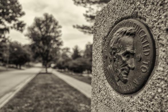 Sepia toned photo of a sign commemorating the old lincoln highway. The concrete sign post has a metal plaque featuring a bust of Abe Lincoln. In the background the highway goes off into the distance.