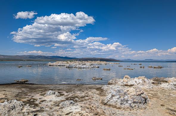 A view over Mono Lake from close to the shore.  Blue sky with white fluffy clouds just above the horizon on the right and billowing up more on the left.  The lake is reflecting the sky and clouds, but isn't perfectly smooth.