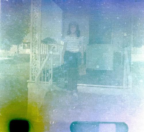 An image taken from the film negative after heavy post-processing reveals a girl standing on the steps of a porch, carrying a bag in each hand. The photo is blurred and contains lots of grain and noise, but is recognizable and even shows some color variations.