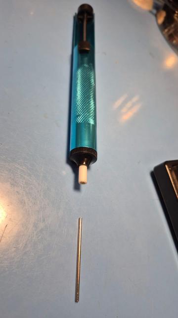 Photo of manual desoldering pump with a thin, round shiny metallic rod next to it, about 1 millimeter in diameter and 40 millimeters in length.