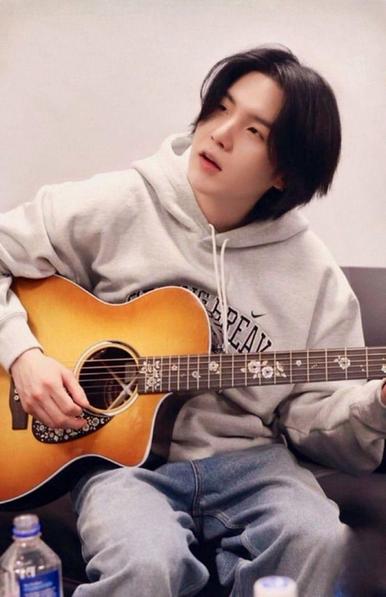 Suga of BTS in a cream hoodie, sitting on a sofa and playing with his D-Day acoustic guitar with the mother of pearl inlay.