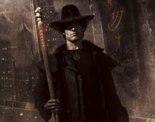 A man wearing a black hat and long black duster coat is holding a large staff. His eyes are hidden in the shade cast by his hat. Behind him is a brown city rainy with tall buildings. He is wearing a 5 pointed star medallion on a chain around his neck. There are glowing sigils or symbols on a wall behind him. There are similar ones on his staff. There's police tape behind him.