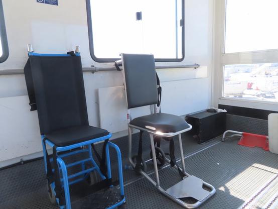 Large metal specialized airplane lift vehicle inside. It has large rectangular windows with rounded corners. There's a small rail underneath the windows for accessibility. Two narrow chairs with no armrests, and black upholstered padding and black woven straps stand against the wall. Some sort of portable barrier with aluminum tubes framing orange construction netting lies on the floor. The end opposite the driving chamber has a larger window, showing us elevated over the airport tarmac, and a view of other planes in the background.