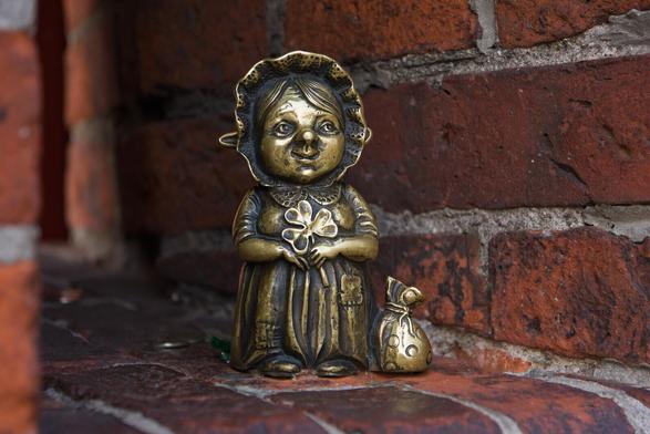 The image depicts a palm-size bronze statue of what appears to be an old woman, standing on the edge of a brick platform. The woman is dressed in clothing reminiscent of old-fashioned European attire for women during colonial times; she wears a bonnet and long sleeves with puffed cuffs typical of that era's fashion. Behind the bonnet ends of two long pointy ears are visible. In her hands, she holds a clover leaf.

Beside the woman stands another bronze object resembling a small sack, possibly representing the simple life associated with early settlers in European colonies. The bricks around these figures are aged red and show signs of wear from weathering over time.