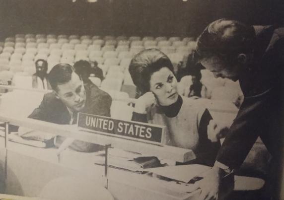Imagen en blanco y negro de una reunión en la ONU, mostrando a tres personas sentadas en una mesa con un cartel que dice 'UNITED STATES'. Dos hombres conversan mientras Shirley Temple escucha atentamente.