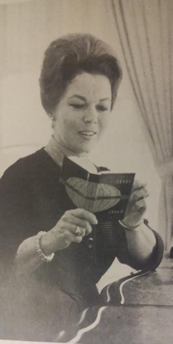 Shirley Temple sosteniendo una tarjeta en una mano mientras lee, con un fondo de cortinas en una habitación.