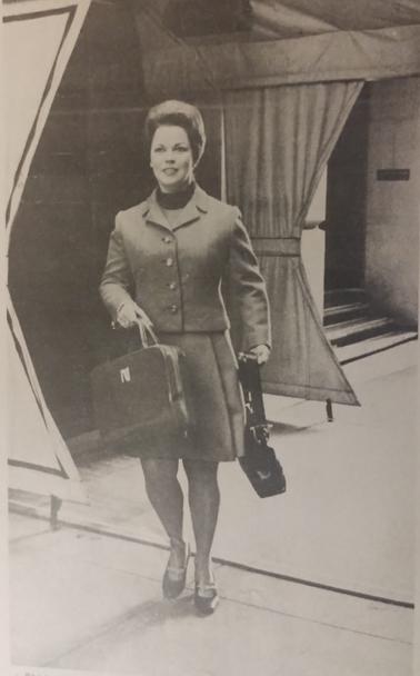 Imagen en blanco y negro de Shirley Temple elegantemente vestida con un traje y tacones, sosteniendo un maletín y una bolsa, caminando hacia la cámara.