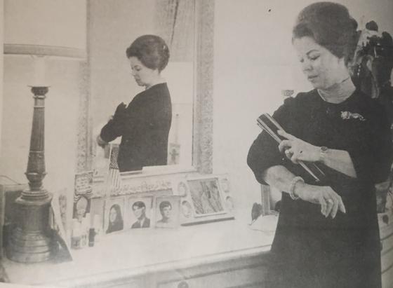 Shirley Temple observándose en un espejo mientras se prepara, con una lámpara y fotos familiares en la mesa frente a ella.