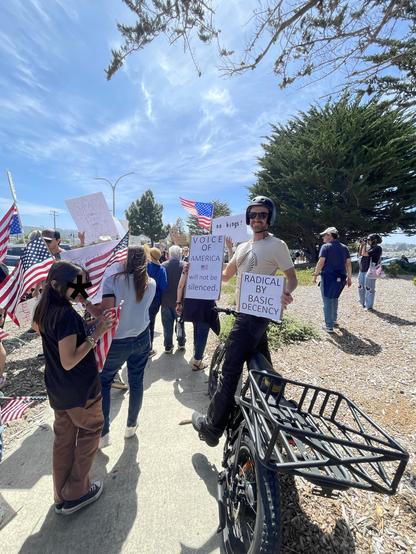 A man on a bicycle has paused to display two signs, which read: “Voice of America will not be silenced” and “Radical by basic decency.” Many U.S. flag-bearers are to his left, including one holding an upside-down flag, the official signal for distress.