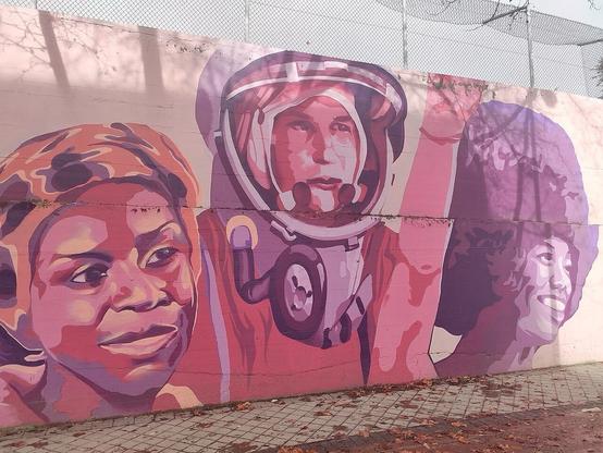 A colorful mural featuring three figures: a smiling girl on the left, an astronaut in the center, and a woman with curly hair on the right. The artwork is characterized by warm, vibrant hues and abstract shapes, set against a textured wall.