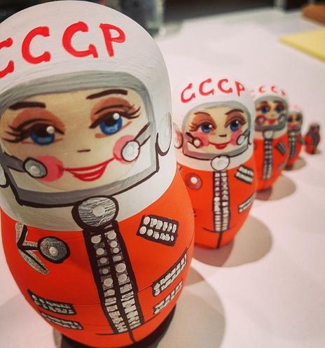 A set of nested Russian dolls (matryoshka) painted to resemble female astronauts in orange jumpsuits, each marked with "CCCP." The dolls feature smiling faces with blue eyes and helmets, arranged in decreasing sizes from front to back.
