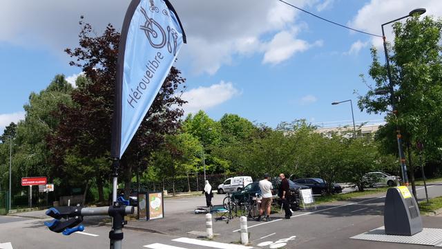 Oriflamme de l'association au premier plan. On voit au loin l'atelier mobile et ses membres installés le long d'une piste cyclable devant un parking voiture.