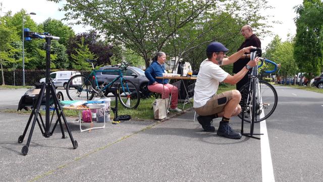 Un membre de l'asso est en train de refixer et régler la selle de son vélo pendant que deux autres membres échangent devant la table de l'atelier.