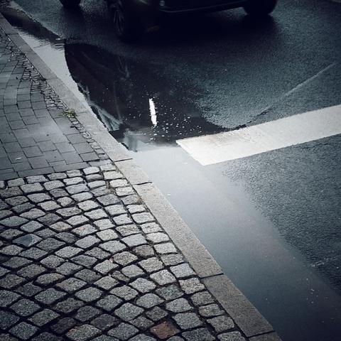 Pfütze am Straßenrand, neben einem Bordstein. Eine weiße Linie führt nach rechts aus dem Bild. Reflektion eines vorüberfahrenden Autos, unscharf. Kleine Pflastersteine auf dem Gehweg, im Vordergrund.