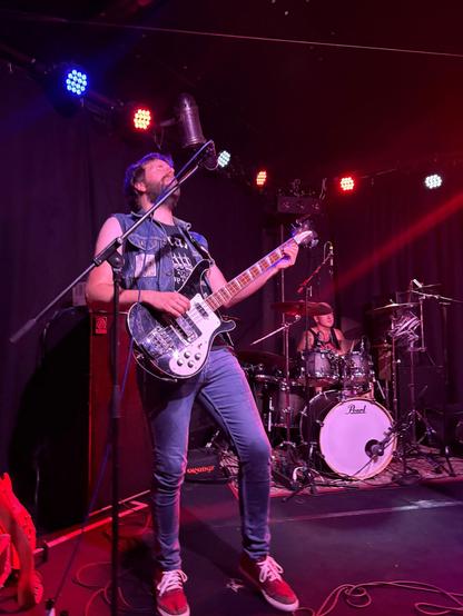 Bearded guy in a jeans jacket plays a Rickenbacker bass on a stage