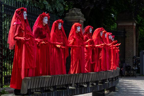 From left to right a diagonal row of men and women standing on a long bench, in long red robes and a red veil over their heads. Their faces are painted white. They hold their hands up in front of them with the palms open and look very serious.