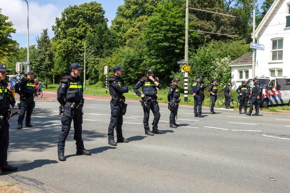 From left to right stands a diagonal line of policemen dressed in black. Behind and in front of them is the street, empty. They stand there relaxed.