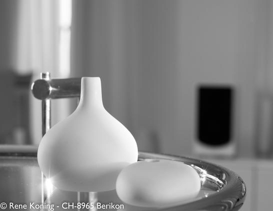 Das Bild zeigt eine minimalistische Schwarz-Weiß-Komposition mit zwei weißen Keramikobjekten (Vasen) auf einer spiegelnden Oberfläche.
Das Objekt auf der linken Seite ist eine Vase mit einem abgerundeten Körper und einem schmalen Hals, während das Objekt auf der rechten Seite ein glattes, ovales Stück ist und fast wie ein grosser, dicker Kieselstein aussieht.
Beide Objekte haben eine matte Oberfläche, was zu der klaren Ästhetik beiträgt. Der Hintergrund ist sanft verschwommen, mit einer Andeutung eines Fensters und einem Lautsprecher, sichtbar. Die spiegelnde Oberfläche unter den Objekten verleiht der Szene einen subtilen Glanz, der das moderne und elegante Gesamtbild unterstreicht.