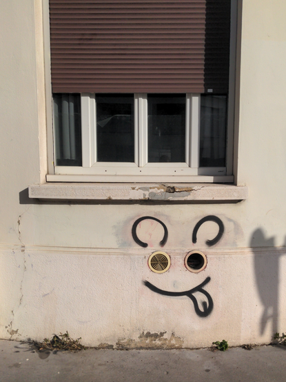 Sous une fenêtre, quelqu'un a ajouté des traits de graf noir autour de deux sorties de tuyau d'aération rond formant ainsi des yeux, et un visage coquin et souriant, tirant la langue, avec des oreilles de souris.