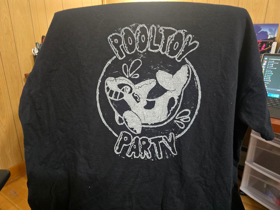 a black shirt with a white block print on it, the print is of a pooltoy orca, above and below it says pooltoy party in inflatable letters, the black part of the orca is white, and the white is black due to the print