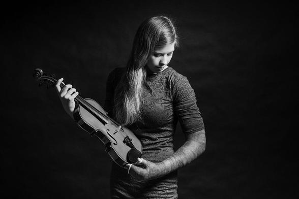 Studioportrait einer jungen Frau, die ihren gesamten linken Arm inklusive Hand und Finger in einem Gips hat und mit der rechten Hand eine Violine hält.
