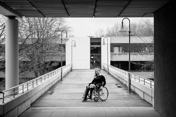 Eine junge Frau sitzt mit zwei Gipsbeinen in einem Rollstuhl auf einer Brücke von einem Wohnhauskomplex zu einem anderen.