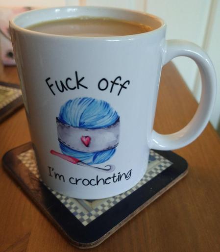 Mug of tea on a bedside cabinet with the words 'fuck off I'm crocheting' surrounding a ball of blue yarn and a crochet hook