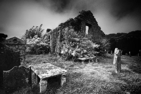 Atmospheric black and white photo of the overgrown gable of a ruined church, with tombstones in front.