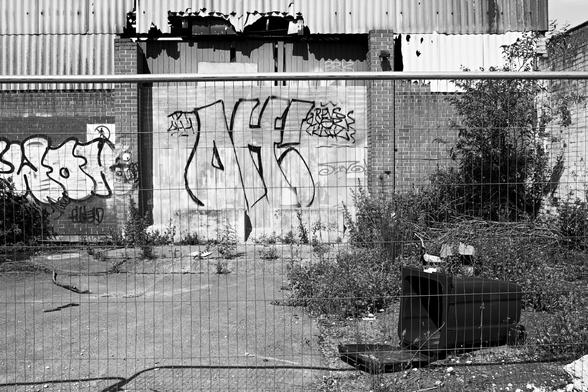 Behind a wire fence, a tipped-over black garbage bin rests on cracked concrete, surrounded by overgrown weeds and scattered debris. The background reveals a derelict industrial building, its facade a patchwork of corrugated metal and brick walls covered in graffiti. Some of the metal sheeting is torn and curling, exposing darker shadows within. Harsh sunlight carves deep contrasts into every surface, highlighting decay and disorder. The image is in black and white, which amplifies the grit and starkness. It's a raw, quiet scene of urban neglect, where discarded infrastructure and untamed nature coexist in the absence of people or repair.