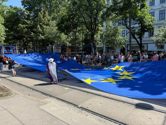 You see a very big EU flag, estimated 8m wide and 20m long carried by two rows of colorful people as part of the pride parade on Ringstraße Wien 14. June 2025.