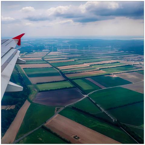 View from an airplane window, part of the wing to the left, a greenish patchwork landscape and a wind farm below.