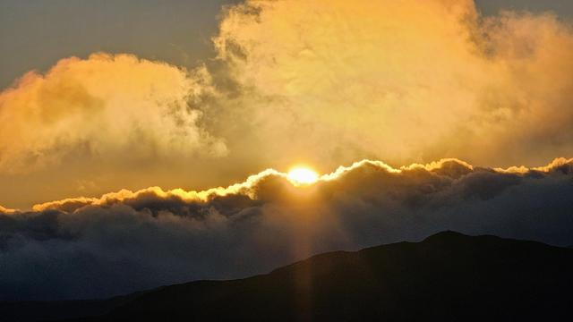 A vibrant sunset captured with the sun bursting through a horizontal band of golden-orange clouds, casting rays of light downwards. Below the clouds, the dark silhouette of a mountain range stretches across the bottom of the frame. The upper portion of the sky is filled with more warm-toned clouds, hinting at the expansive and beautiful skyscape.