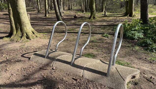 In a sunny mature woodland, three wavey concrete blocks hold unusually shaped steel cycle racks - emerging at an angle from the concrete in a soft, rounding form. Decaying text applique reads ‘Law Cycles’ with a local phone number gradually fading away with time.