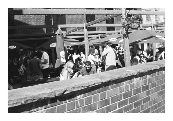 This is a black and white photograph of an outdoor gathering, likely a pub or restaurant patio, bustling with people. In the foreground, a low brick wall runs horizontally across the bottom third of the image, separating the viewer from the scene. Behind the wall, numerous individuals are visible, some seated at tables and others standing and socialising. There are several large, open umbrellas providing shade, and what appears to be a wooden or metal pergola structure overhead. The background features a brick building, suggesting an urban setting. The overall impression is one of a lively, social atmosphere.