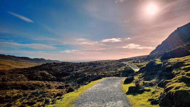 A wide-angle landscape shot of a winding gravel path leading through a rugged, moss-covered landscape under a dramatic sky. The sun, bright and slightly hazy, is positioned in the upper right, casting a warm glow over the scene. The path curves gently to the right, with rocky, dark terrain on the left and a steep, sun-drenched, mossy hillside on the right. In the distance, a hint of the sea is visible under a vast sky with soft clouds.