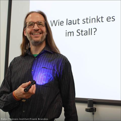Mann mit Brille und langem Haar lächelt vor einer Präsentationswand mit dem Text 'Wie laut stinkt es im Stall?'