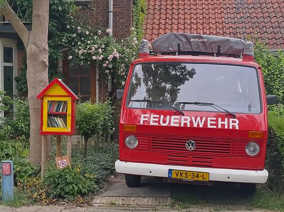 Rood met gele minibieb in een tuin naast een oude brandweerbus (met opschrift Feuerwehr) die nu als camper in gebruik is.