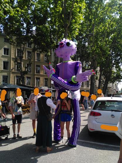 Photo de rue avec une grande marionnette de 3m de haut. Sa tête est en forme de poisson-globe, elle porte un survêtement violet et pose façon Jul. Une personne placée en dessous utilise des perches pour bouger ses bras.