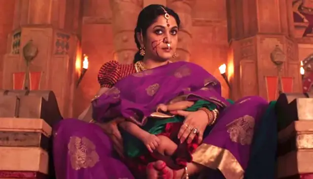 A richly bejeweled woman dressed in a finely woven sari with gold trim, sitting in lotus pose on a stone throne, with two babies on her lap, each one breastfeeding (they're covered by her sari but the pose is clear). There's a large streak of blood on her face.
