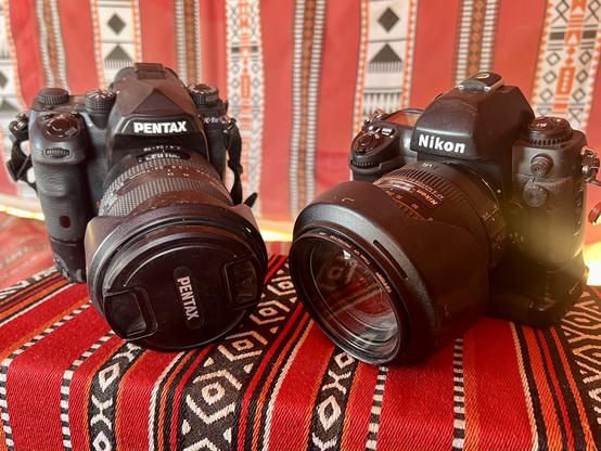 A large Pentax and a large Nikon camera sitting side by side on a red fabric cushion.