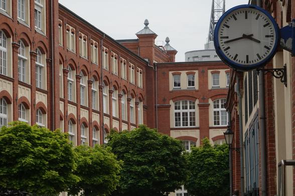 Backsteingebäude, Grünzeug und große Analoguhr
Im Hintergrund ragt ein runder Büroturm über das Dach