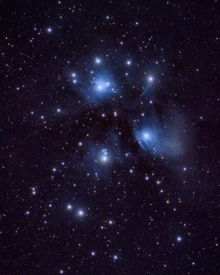 An astrophotography shot taken from Bubulcus & Bolotas revealing the captivating beauty of the Pleiades, or Seven Sisters. Intense blue stars glow brightly, scattering their light through a wispy blue reflection nebula that appears to swirl around them in the vastness of space.
